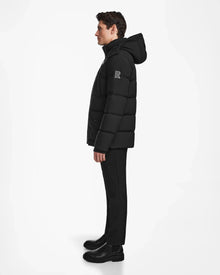 A man stands in profile against a white background, wearing the Rudsak CARLOS - M124509 Black quilted parka coat with a hood, paired with black pants and boots.