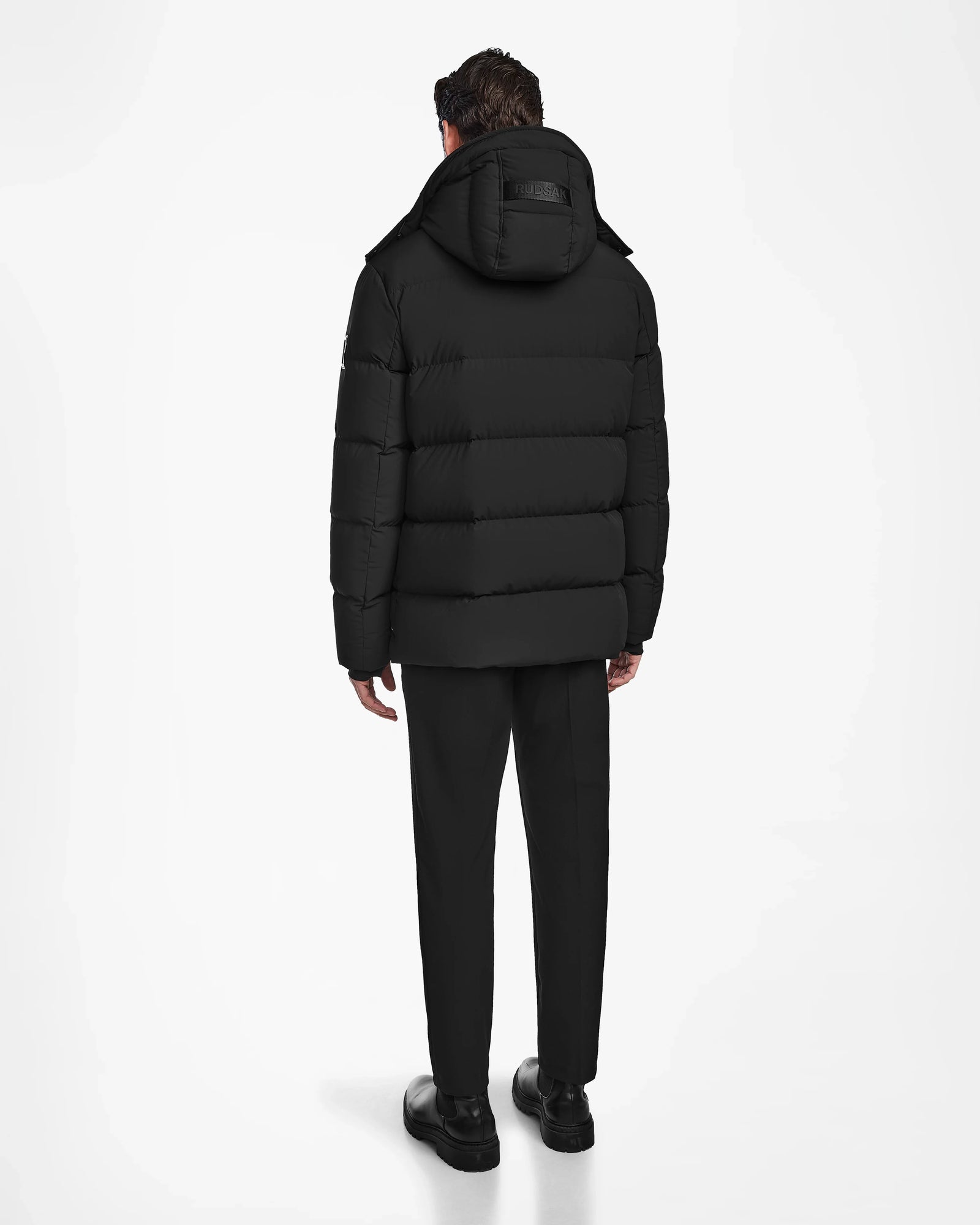 A man stands facing away, wearing the Rudsak CARLOS - M124509 Black men’s down parka with a hood, along with black pants and shoes, against a plain white background.