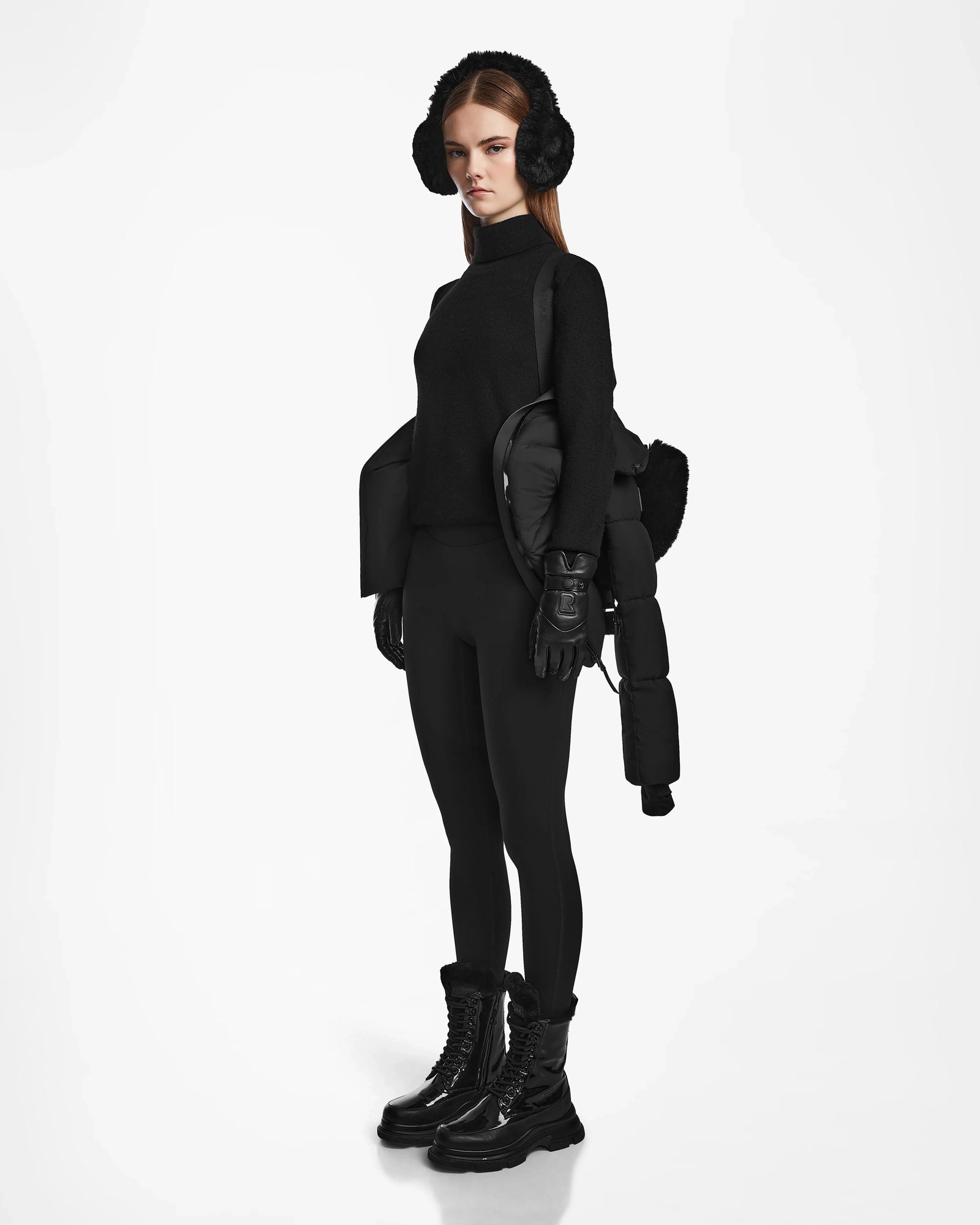 A woman in an all-black winter outfit stands against a plain white background, carrying the Rudsak CHRISTELLE M - L123525 Black women’s quilted down puffer over her arm.