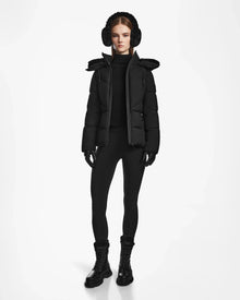 A person stands facing forward against a white background, wearing the Rudsak CHRISTELLE M - L123525 Black sustainable winter coat with a fur-trimmed hood, black earmuffs and gloves, leggings, and lace-up boots.