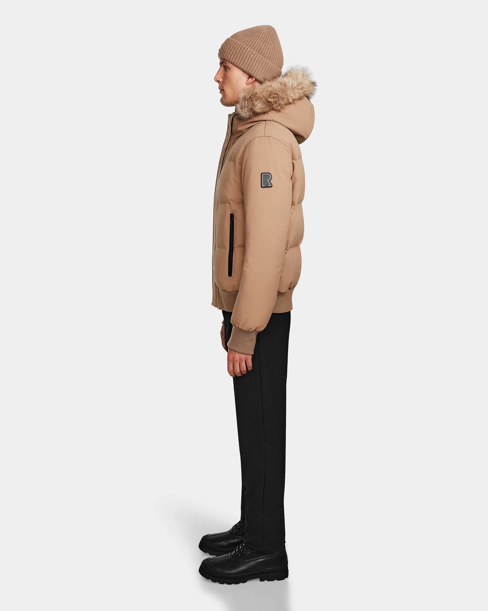 A person stands in profile wearing the Rudsak DALLAS - M122501 Sand weather-resistant bomber jacket with RDS-certified down, paired with black pants, black shoes, and a beige knit beanie against a plain white background.