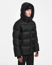A person wearing the Rudsak DANIEL M - M124510 Black mens hooded down puffer jacket and a black beanie stands against a plain white background.
