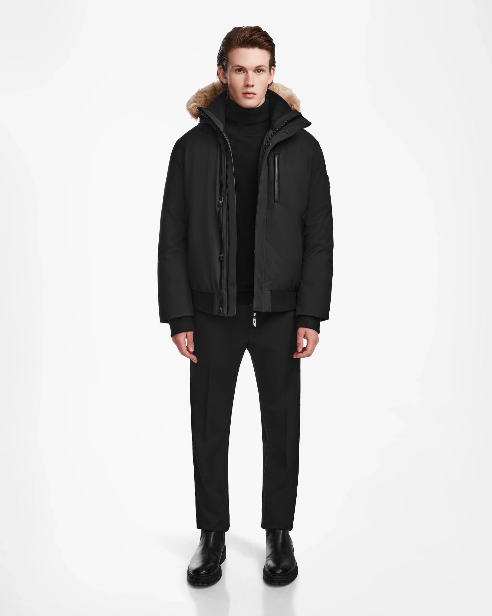 A man faces forward against a white background, wearing the Rudsak DEACON - M123513 Black/Natural bomber-style down puffer with recycled leather trim and RDS-certified down, paired with a black turtleneck, pants, and boots. His hands rest at his sides.
