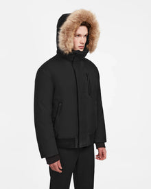 A person wears the Rudsak DEACON - M123513 Black/Natural men's winter coat, a black bomber down puffer with recycled leather trim and a fur-lined hood, featuring a zippered chest pocket, against a plain white background with matching black pants.