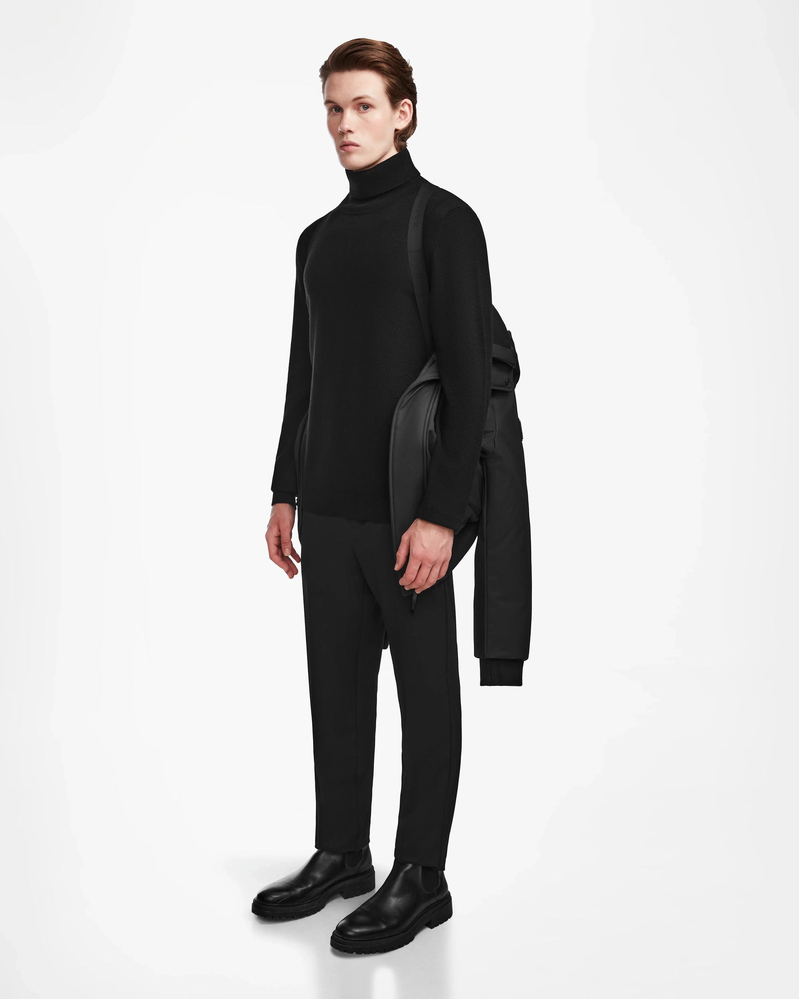 A man stands against a white background wearing black clothes and holding the Rudsak DEACON - M123513 Black/Natural bomber-style down puffer with recycled leather trim over his left shoulder.