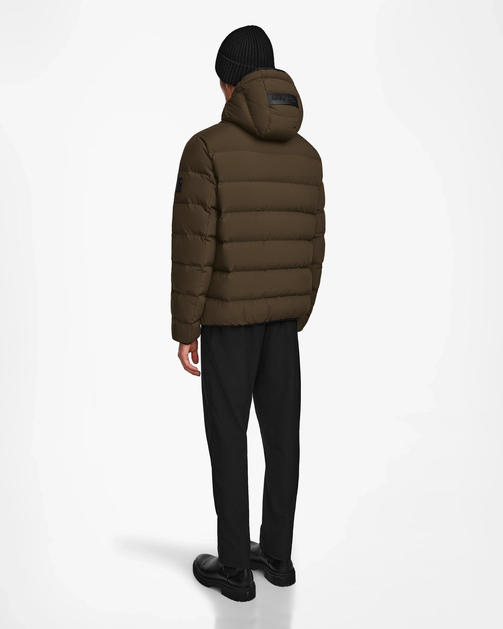 A person stands with their back to the camera, wearing the Rudsak DEMIAN M - M123507 Army mens down puffer jacket, paired with black pants, shoes, and a beanie against a plain white background.