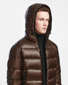 A young person wears the Rudsak DEMIAN S - M123508 men’s shiny espresso down puffer jacket with hood up, looking slightly to the side against a plain white background.