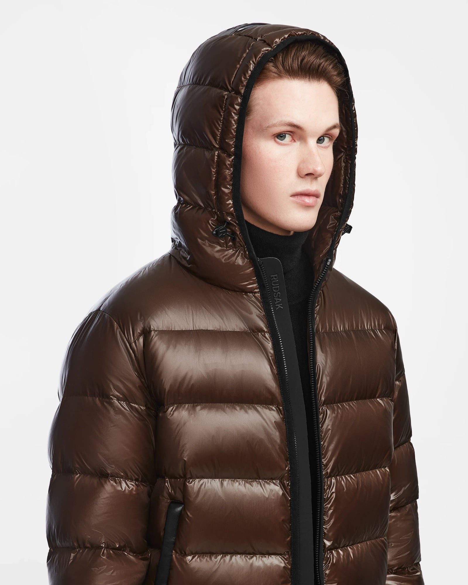 A young person wears the Rudsak DEMIAN S - M123508 men’s shiny espresso down puffer jacket with hood up, looking slightly to the side against a plain white background.