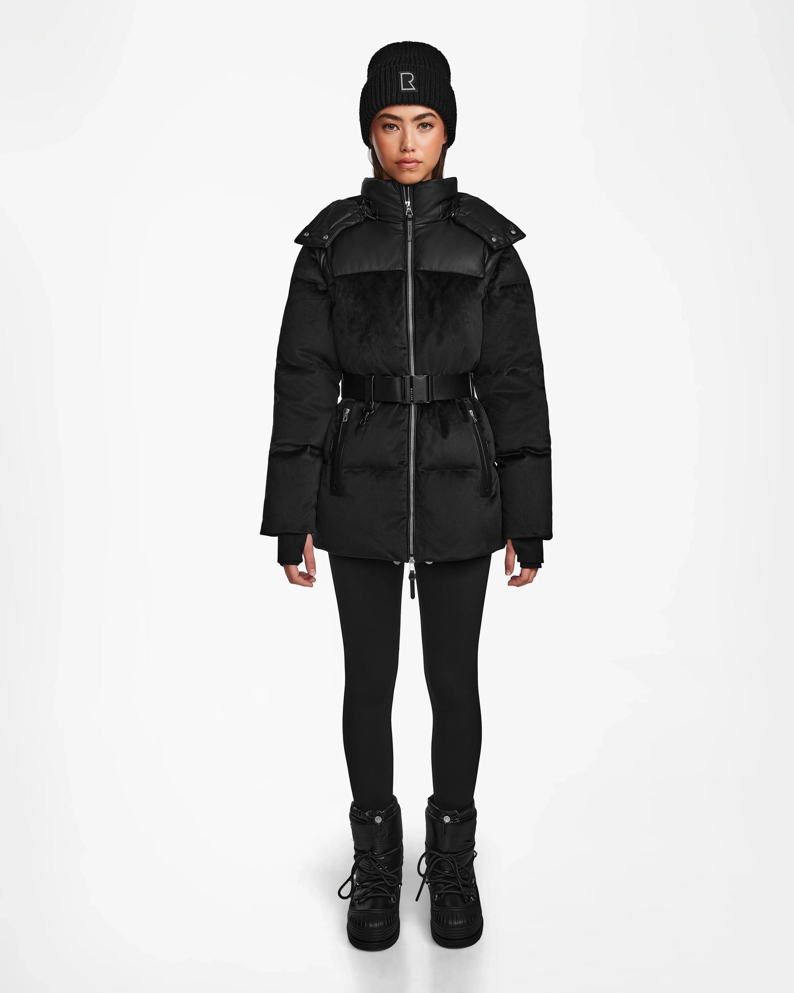 A woman faces forward against a plain white background, wearing the Rudsak DONIA - L124563 Black belted womens down puffer coat, paired with a black beanie, black leggings, and black winter boots.