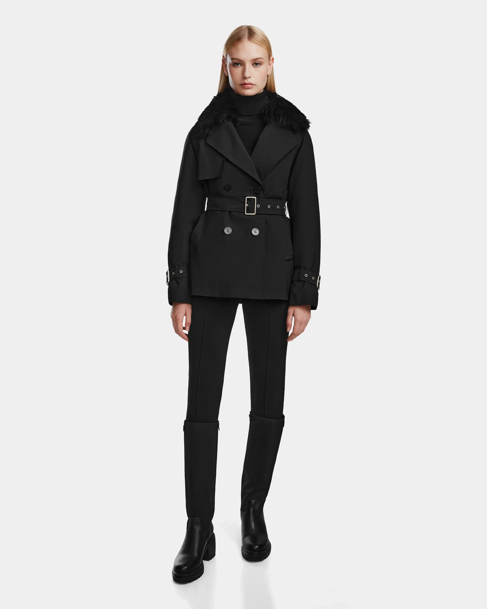 A woman stands facing forward against a plain white background, wearing the Rudsak ELENOR - 8124707 Black womens trench coat with a wide shearling collar, paired with black fitted pants and black ankle boots.
