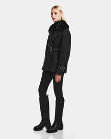 A woman stands slightly sideways against a plain light background, wearing the Rudsak ELENOR - 8124707 Black trench coat with a shearling collar, paired with black pants and tall black boots.
