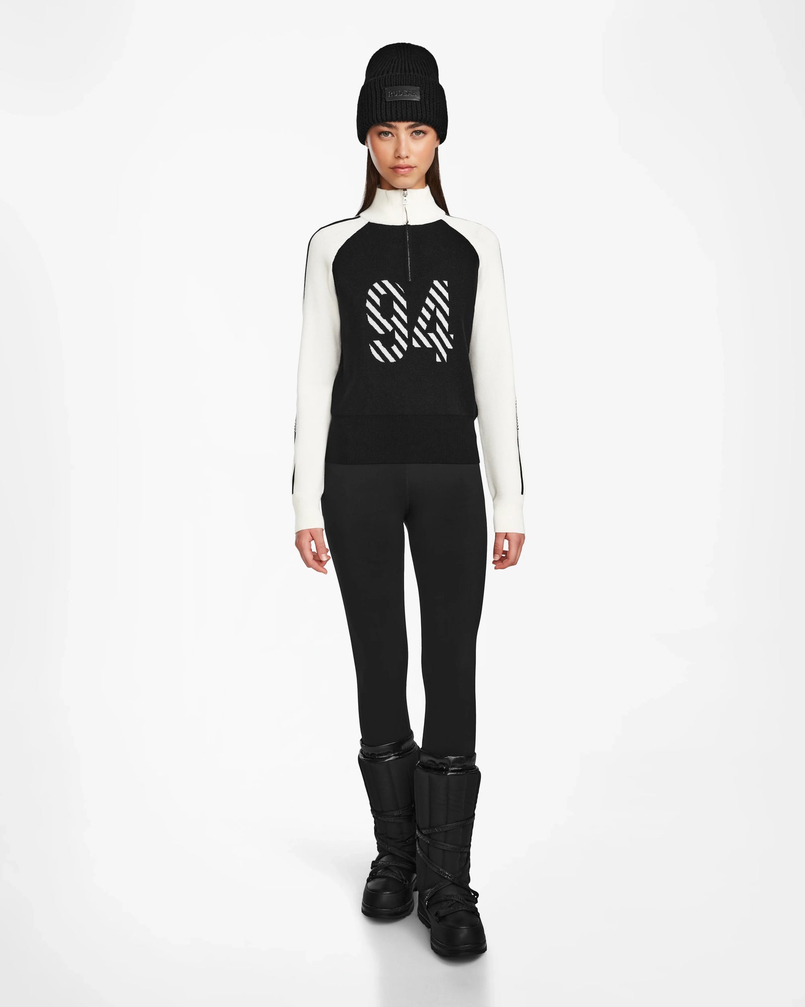 A woman faces forward against a white background, dressed in Rudsak’s FANNY - 8524506 Black après-ski sweater with “84” on the front, black leggings, snow boots, and a knit beanie—ideal for winter layering.
