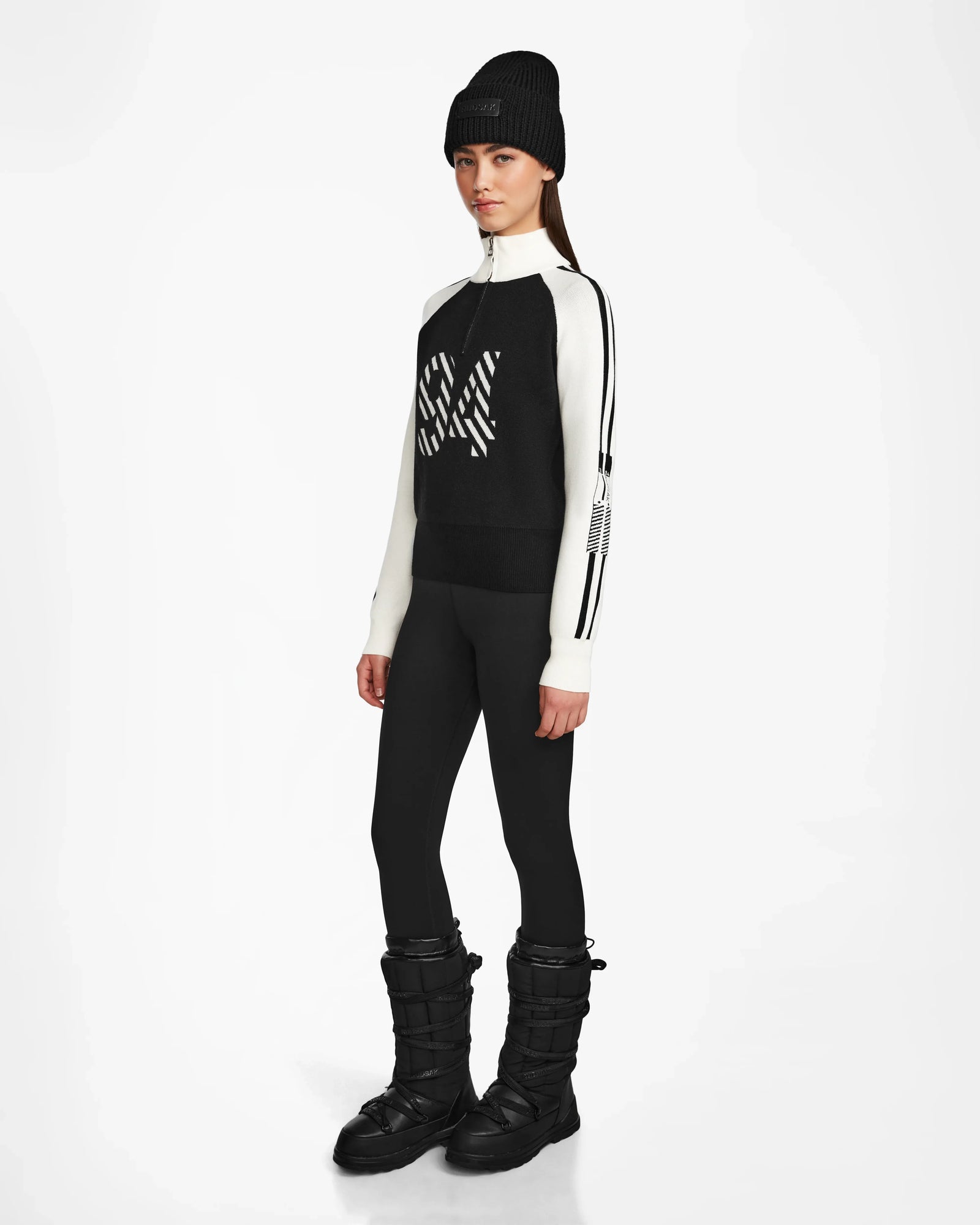 A woman stands against a white background wearing the Rudsak FANNY - 8524506 Black beanie, an après-ski sweater, black leggings, and snow boots. She faces forward with her arms at her sides.