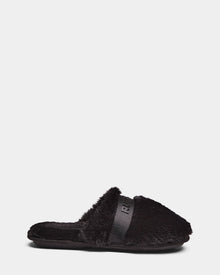 The Rudsak FLUFF - L223509 Black is a cozy black faux-fur slipper with an open back, flat sole, and an embossed strap across the top, shown against a plain white background.