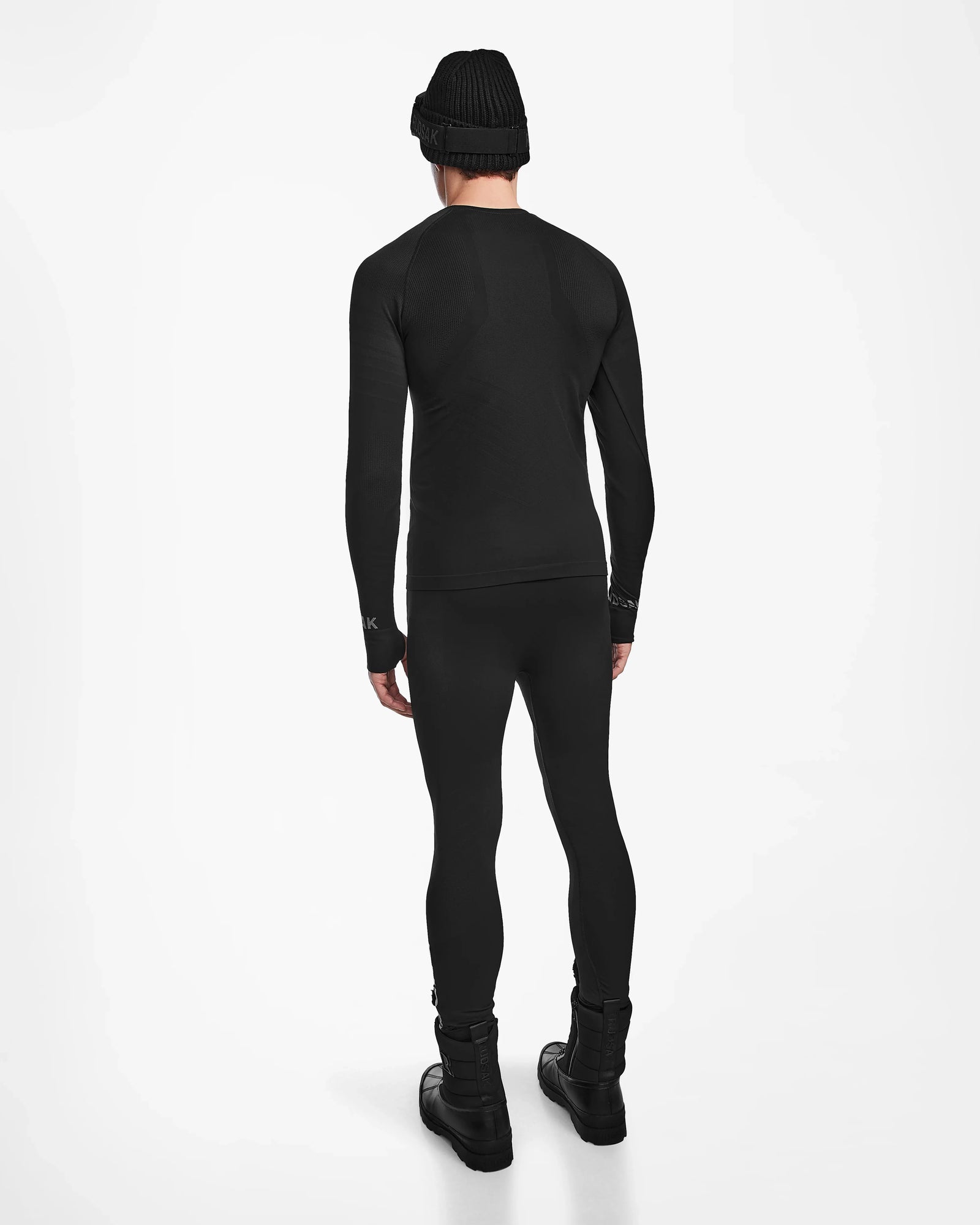 A person showcases the back view of the Rudsak FRANC - 6524541 Black ski underlayer outfit against a plain white background, wearing a black beanie, long-sleeve top, leggings, and boots.