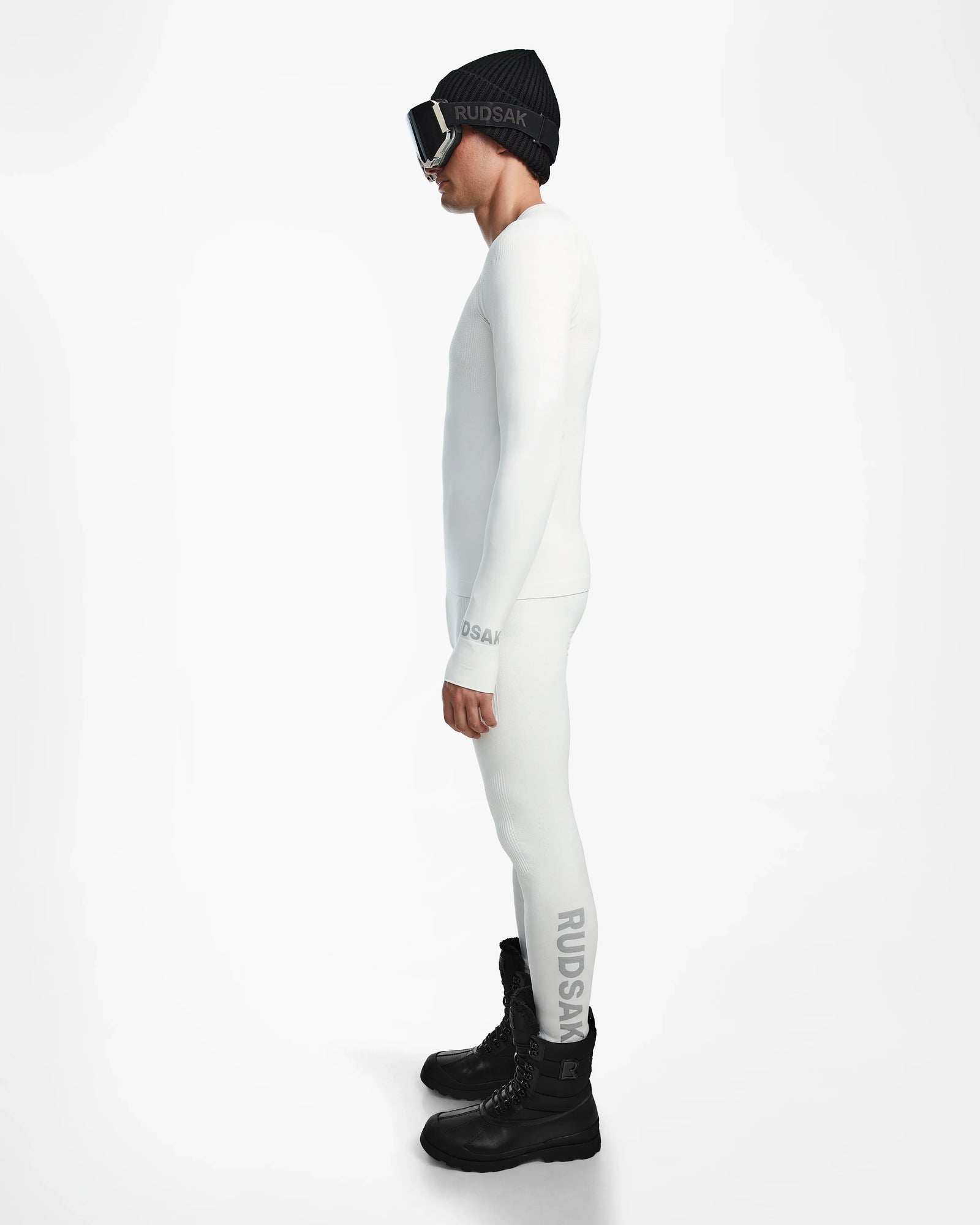 A man in a white Rudsak FRANC - 6524541 Iceland stretch base layer stands in profile, wearing black winter boots, a black beanie, and ski goggles—ready for action against a plain white background.