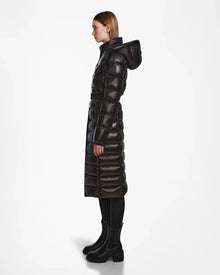 A woman in profile models the Rudsak FRIDA L-S - L124525 Black, a shiny quilted maxi down puffer with hood, paired with black leggings and chunky boots—a chic winter statement—against a plain white background.