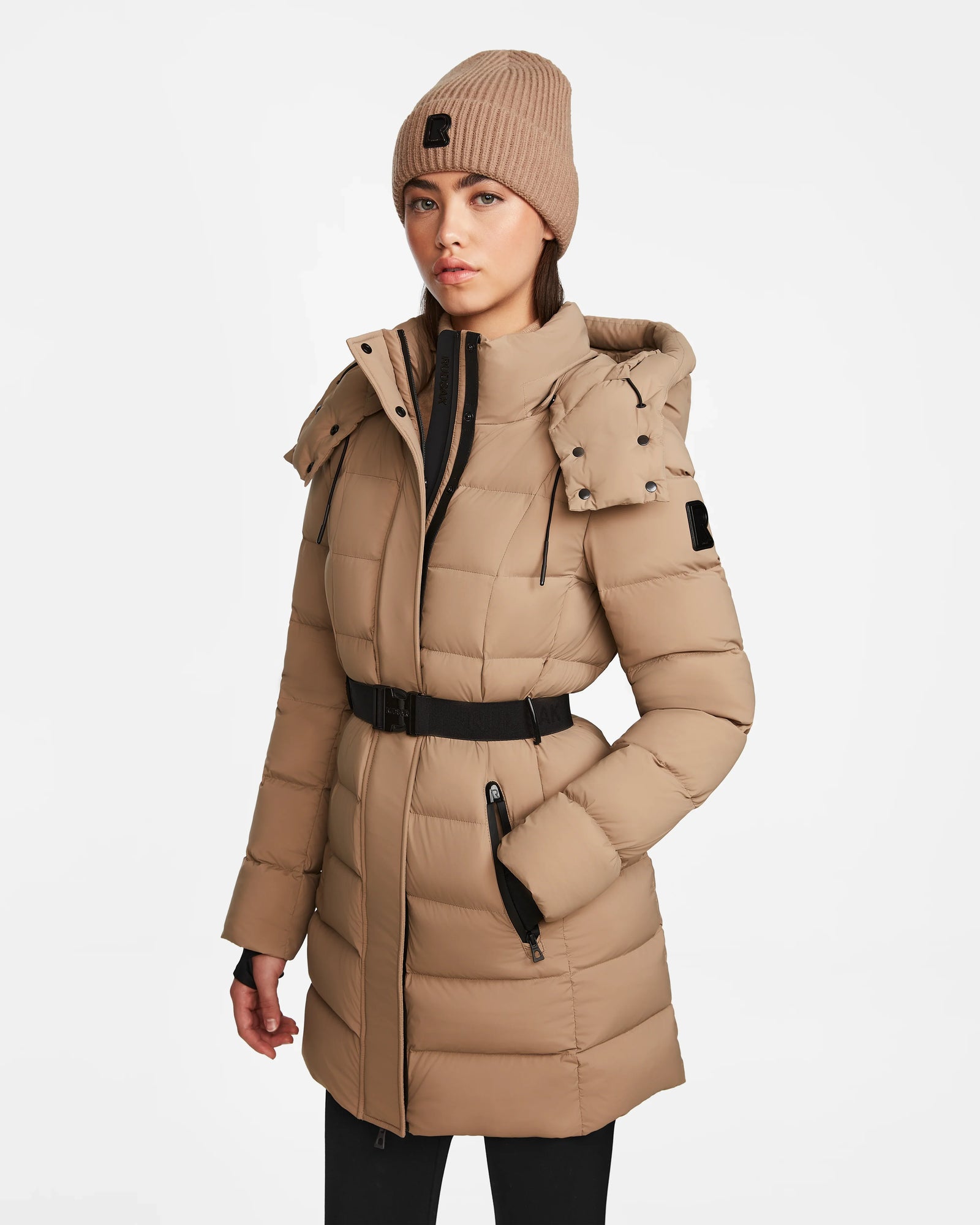 A woman wearing the Rudsak FRIDA M - L124524 Sand down jacket with a belt, black zippers, removable hood, and a matching beige knit beanie stands against a plain white background.