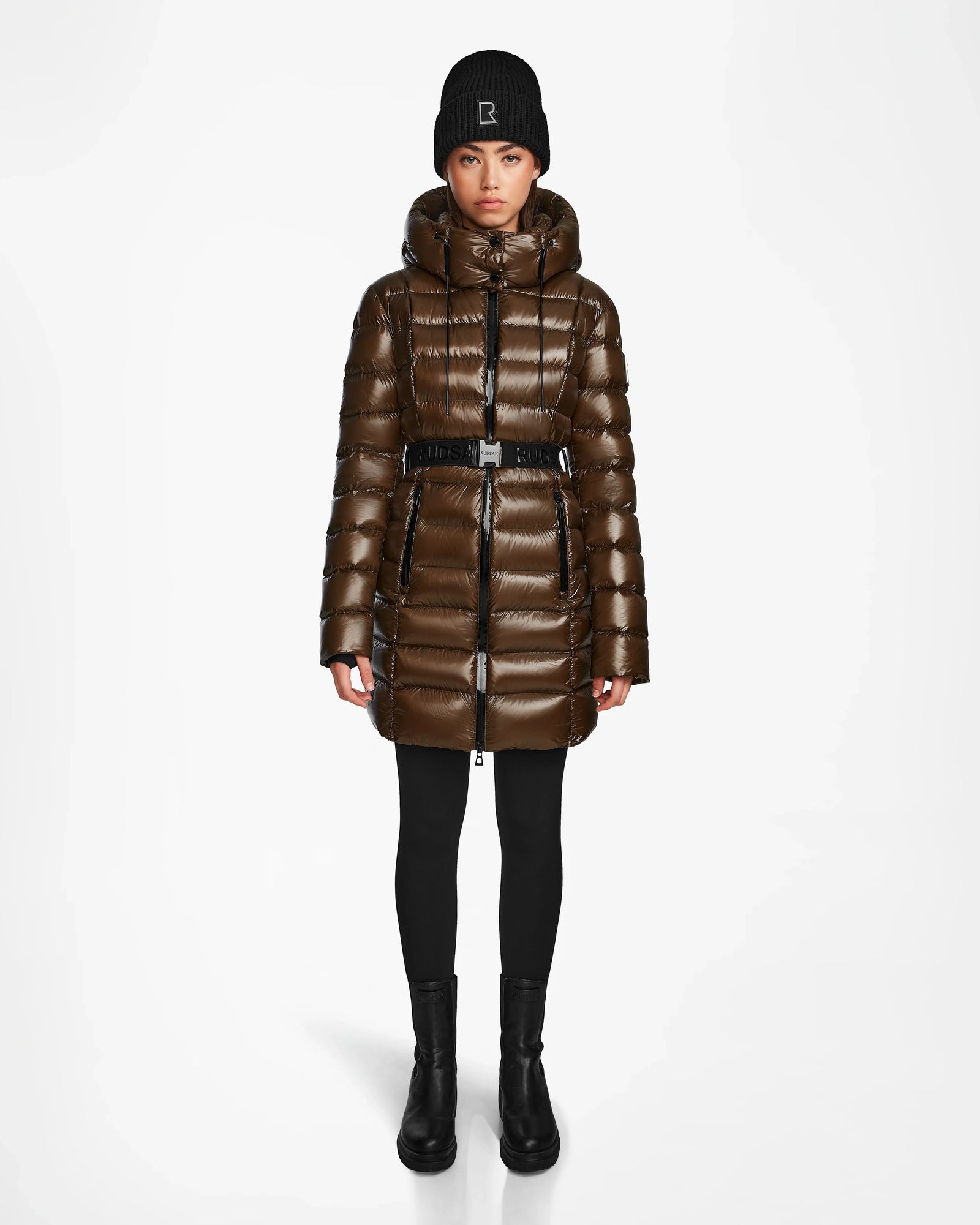 A person stands facing forward wearing the Rudsak FRIDA S - L123513 women’s mid-length down jacket in light olive with a removable hood, black leggings and boots, and a black knit beanie on a plain white background.