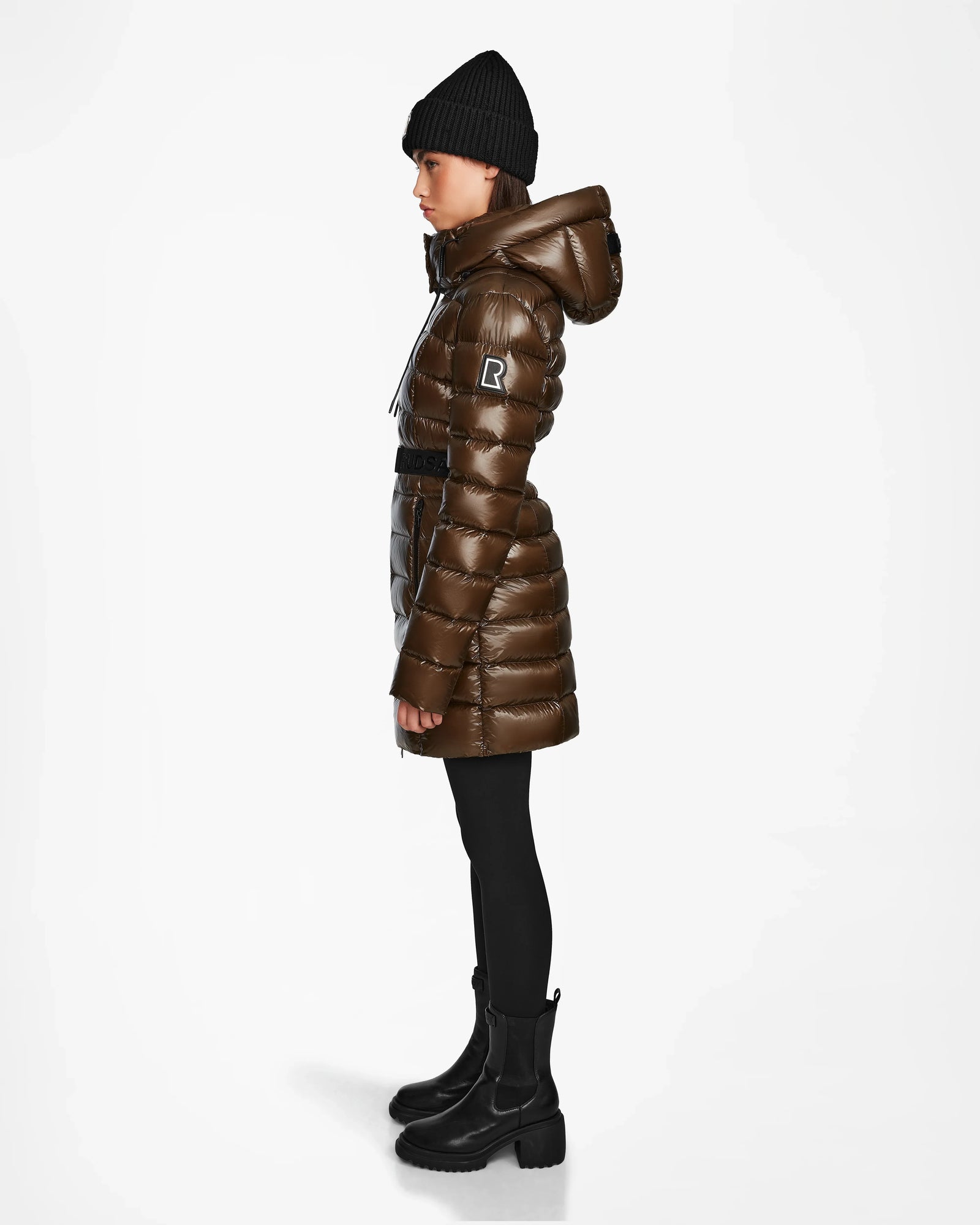 A person stands in profile wearing the Rudsak FRIDA S - L123513 Light Olive women's mid-length down jacket with a hood, black knit beanie, black leggings, and black boots against a plain white background.