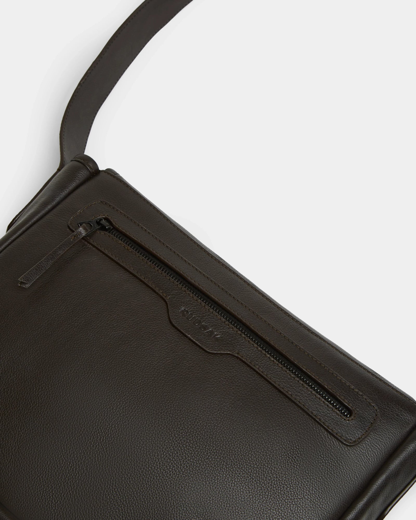 Close-up of the Rudsak GRAYSON - 8324512 Espresso crossbody bag, featuring dark brown textured leather, a front zipper pocket, an adjustable shoulder strap, and the subtly embossed brand name above the zipper for added refinement.