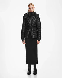 A woman stands facing forward wearing the Rudsak HANNA X - 8123558 Black luxury down jacket, paired with a long black skirt and black boots, against a plain white background.