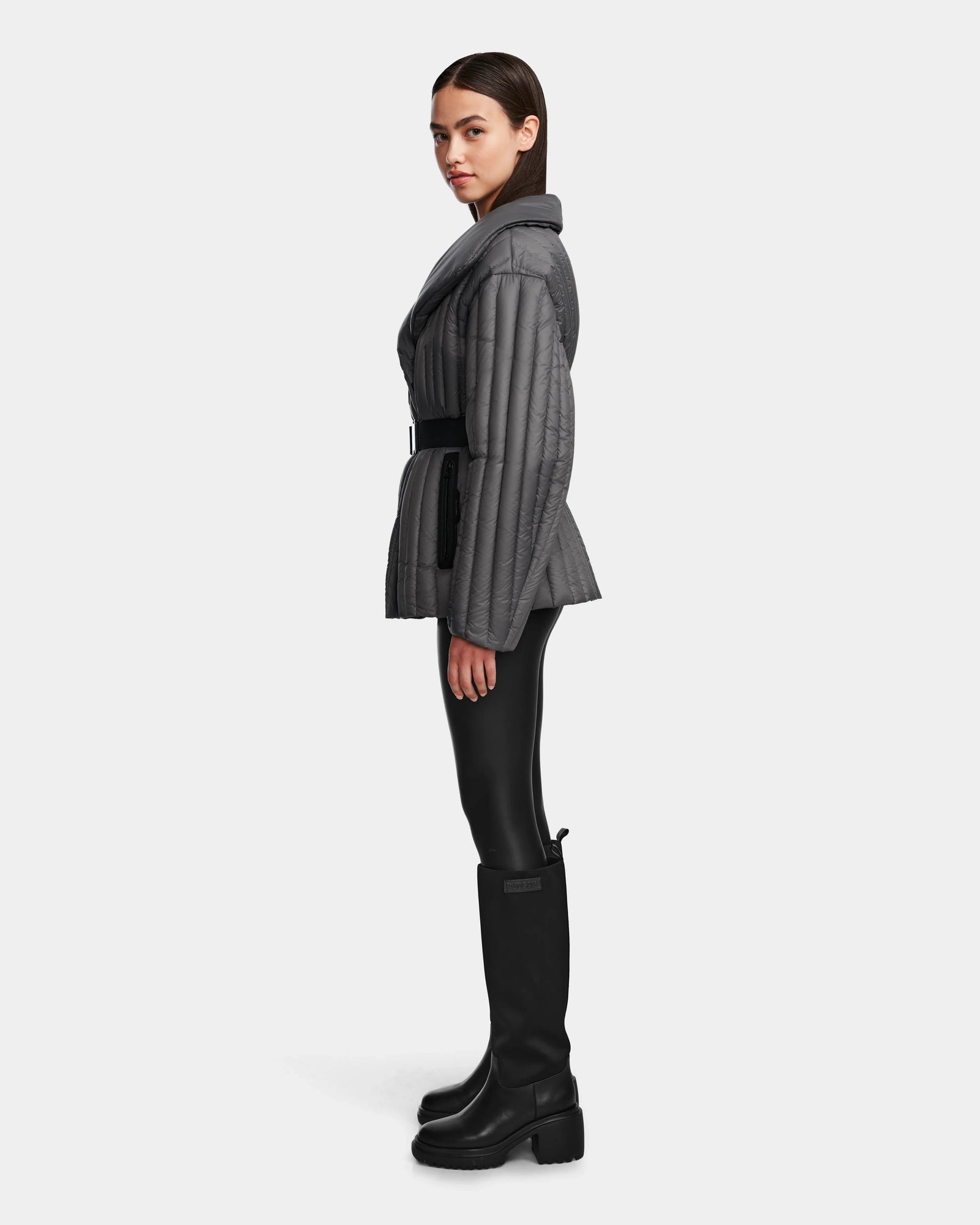 A woman in profile wears the Rudsak INES T - 8124625 Grey quilted jacket with a belt, paired with black leggings and tall black boots, set against a plain white background.