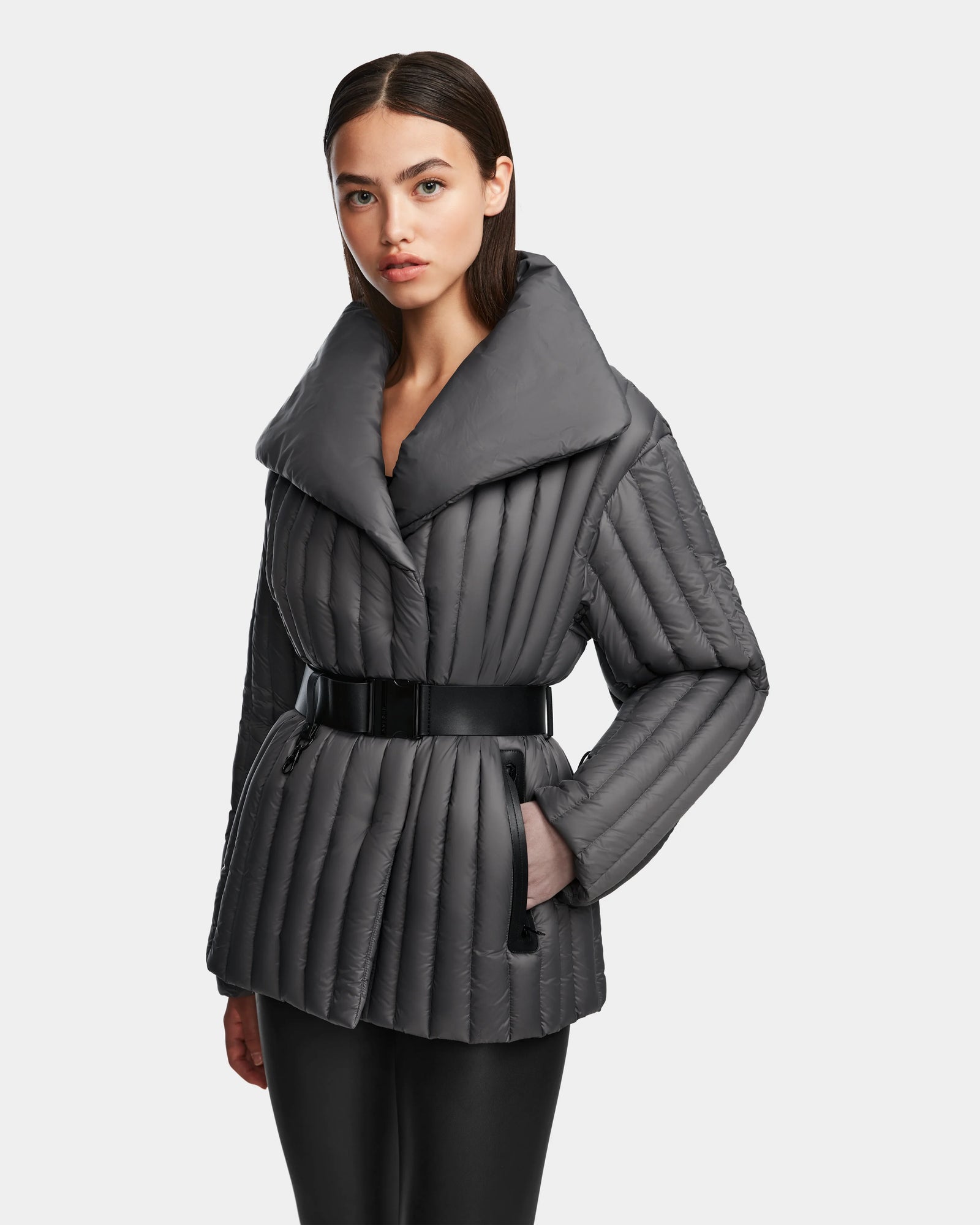 A woman wears the Rudsak INES T - 8124625 Grey RDS certified down quilted jacket with a wide collar and black belt, standing with one hand in her pocket against a plain light background.