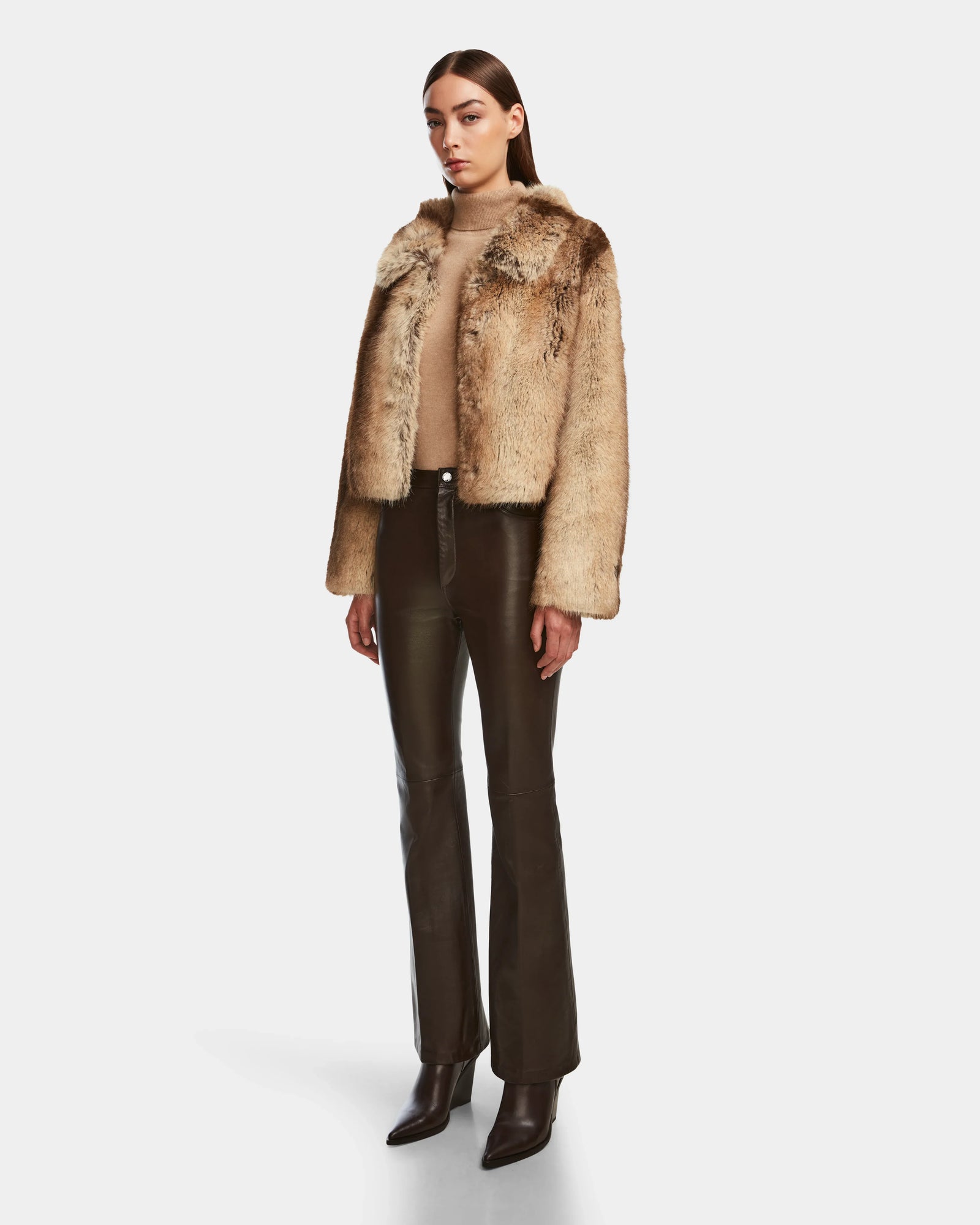 A woman wears the Rudsak JOSILYN - 8124792 Mocha faux fur jacket over a beige top, paired with dark brown leather pants and pointed-toe boots, all set against a plain white background.