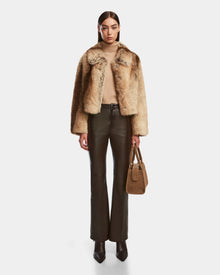 A woman faces forward against a plain white background, wearing the Rudsak JOSILYN - 8124792 Mocha plush-texture women’s faux fur jacket, beige turtleneck sweater, dark brown leather pants, pointed black boots, and holding a brown suede handbag.