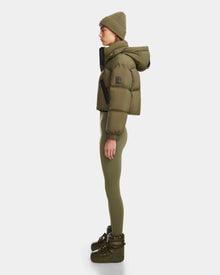 A person stands in profile wearing the Rudsak KARLEE - 8124721 cropped down puffer in Loden Green, paired with a matching beanie, fitted leggings, and platform boots against a plain white background.