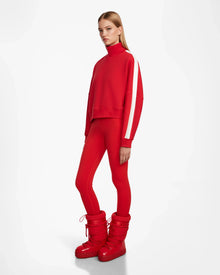 A woman with straight blonde hair wears the Rudsak KELLIE - 8524615 Flame Red mock neck top with white sleeve stripes, paired with red leggings and red padded boots, standing against a plain white background.