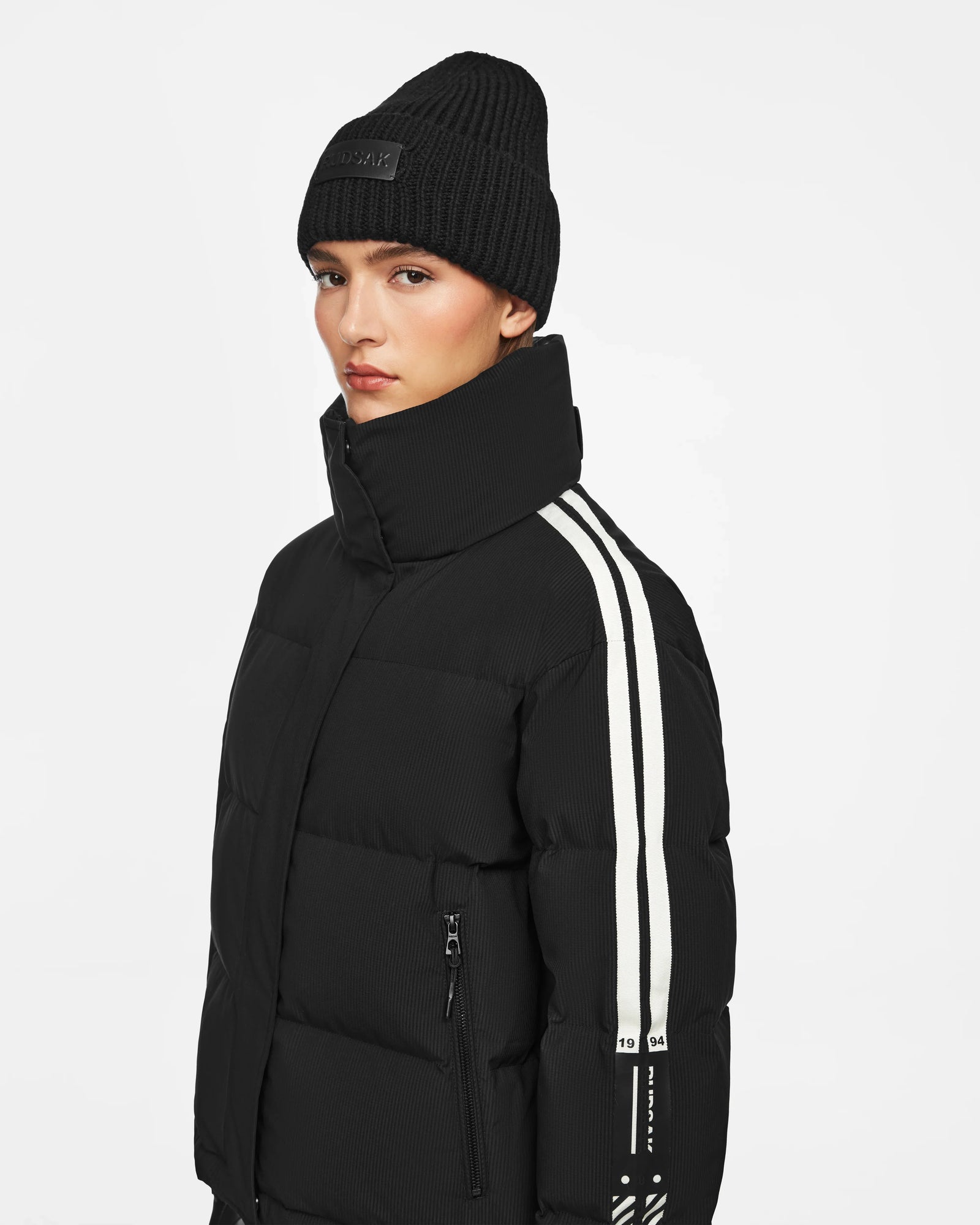 A person in the Rudsak KIMIA - 8124716 Black women’s ski jacket, featuring white sleeve stripes, and a black knit beanie, stands against a plain white background.