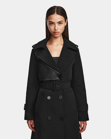 A woman with straight dark hair wears the Rudsak KINDA - 8124690 Black trench coat, featuring leather details, a belted waist, and double-breasted buttons, standing against a plain white background.