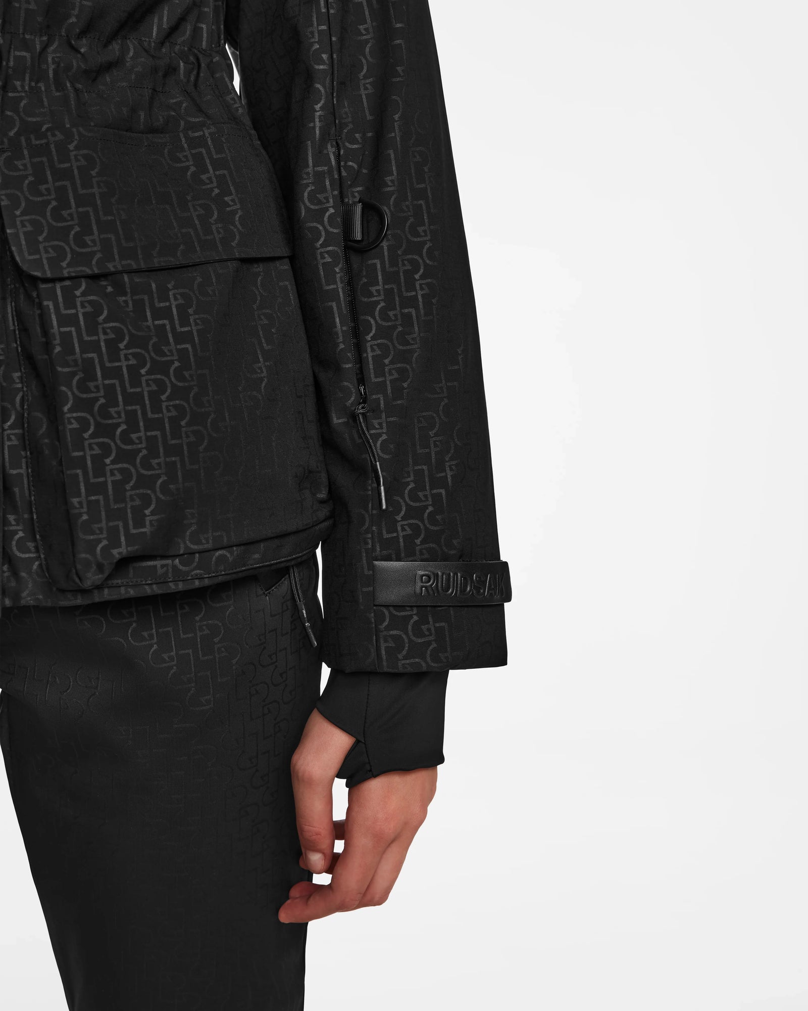 A person wears the Rudsak LUX MO - 8124570 Monogram Print Black womens ski jacket and pants, featuring a subtle repeating pattern. The left arm shows RUNCL on the jacket sleeve strap. The background is plain white.