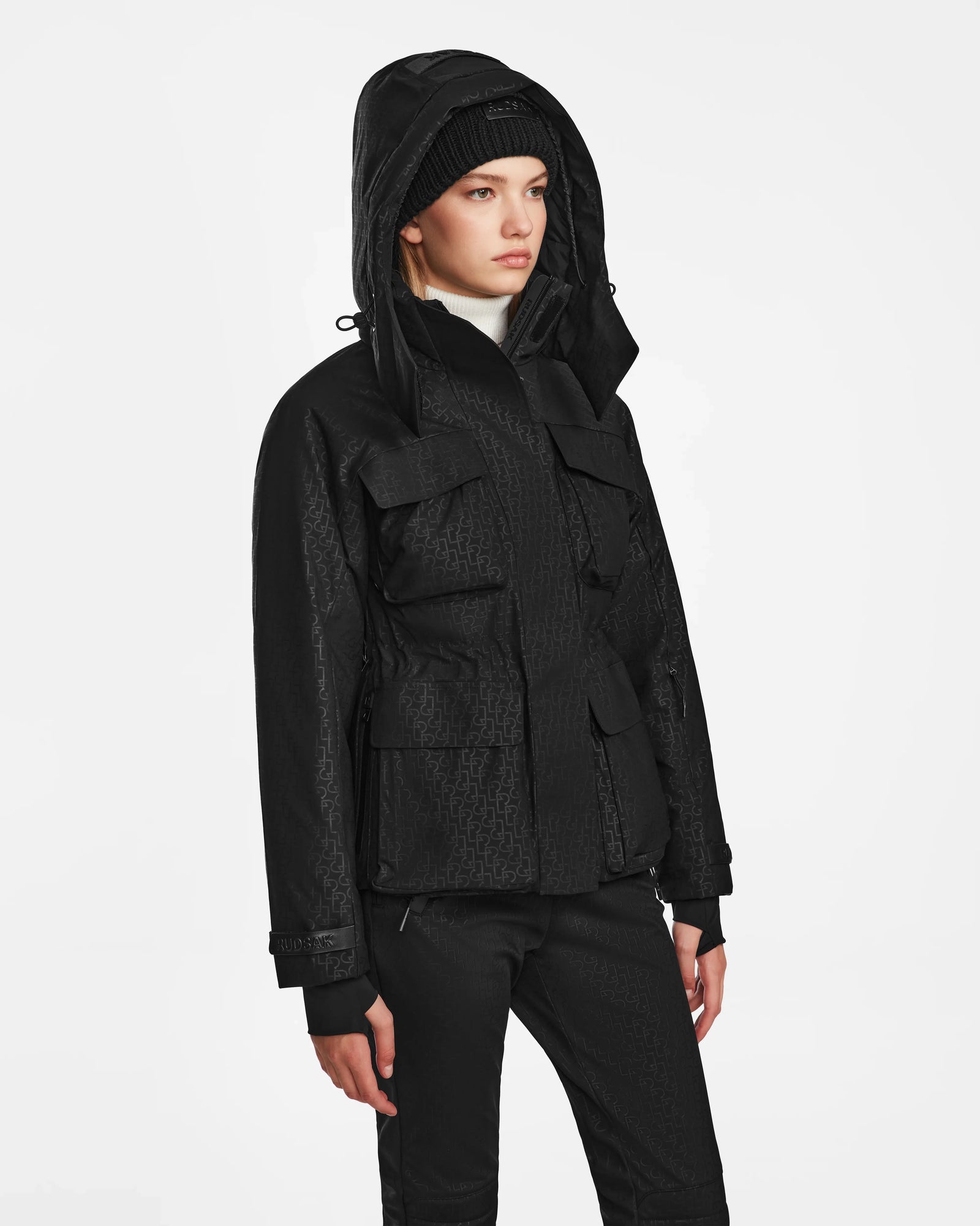 A person wearing the Rudsak LUX MO - 8124570 Monogram Print Black ski jacket, paired with black pants and a black beanie, stands against a plain white background, gazing slightly to the side with a neutral expression.