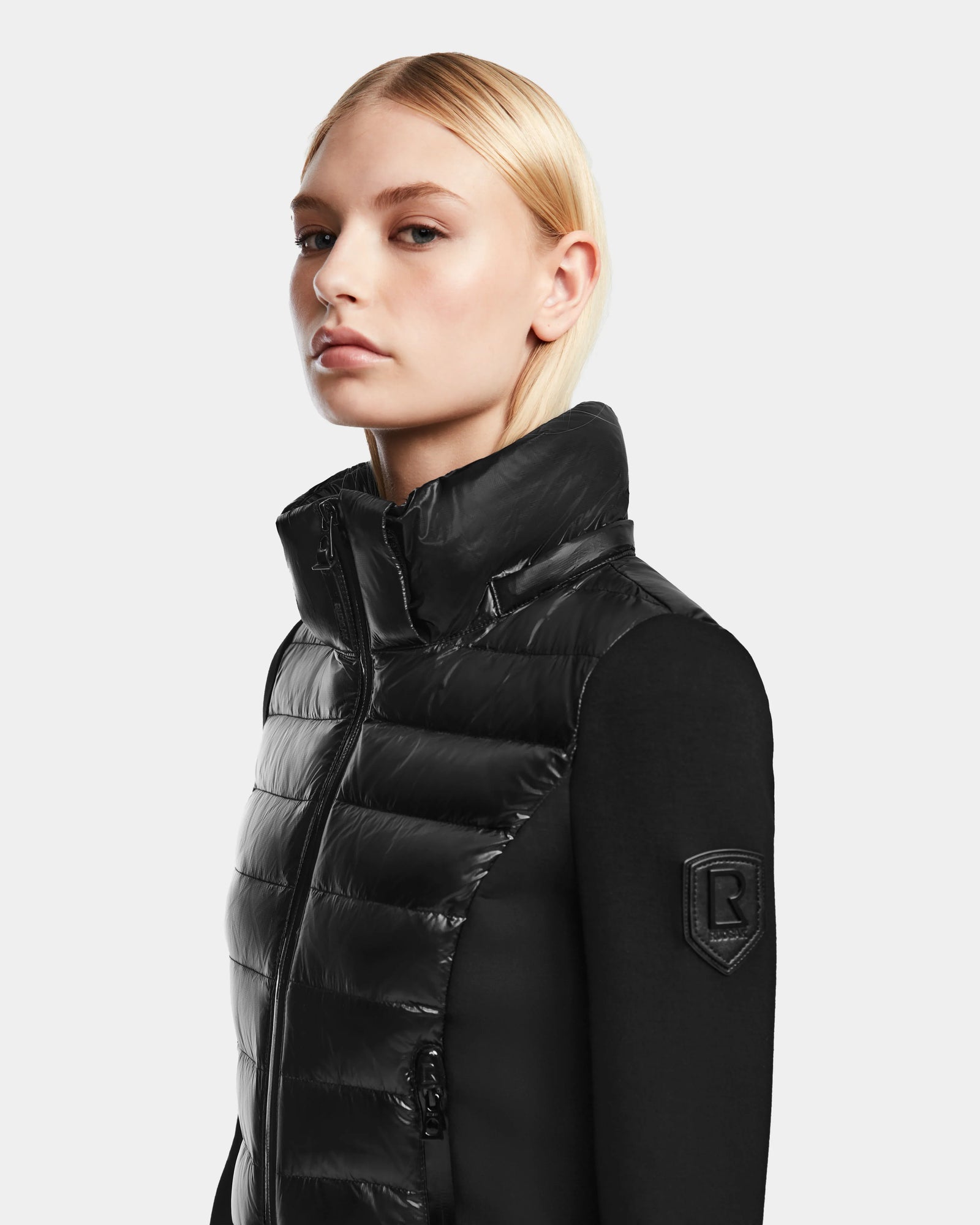 A woman with straight blonde hair models the RUDSAK MABS - 8122020 BLACK womens puffer jacket, featuring a high collar, sleeve patch, and zip, against a plain light gray background.