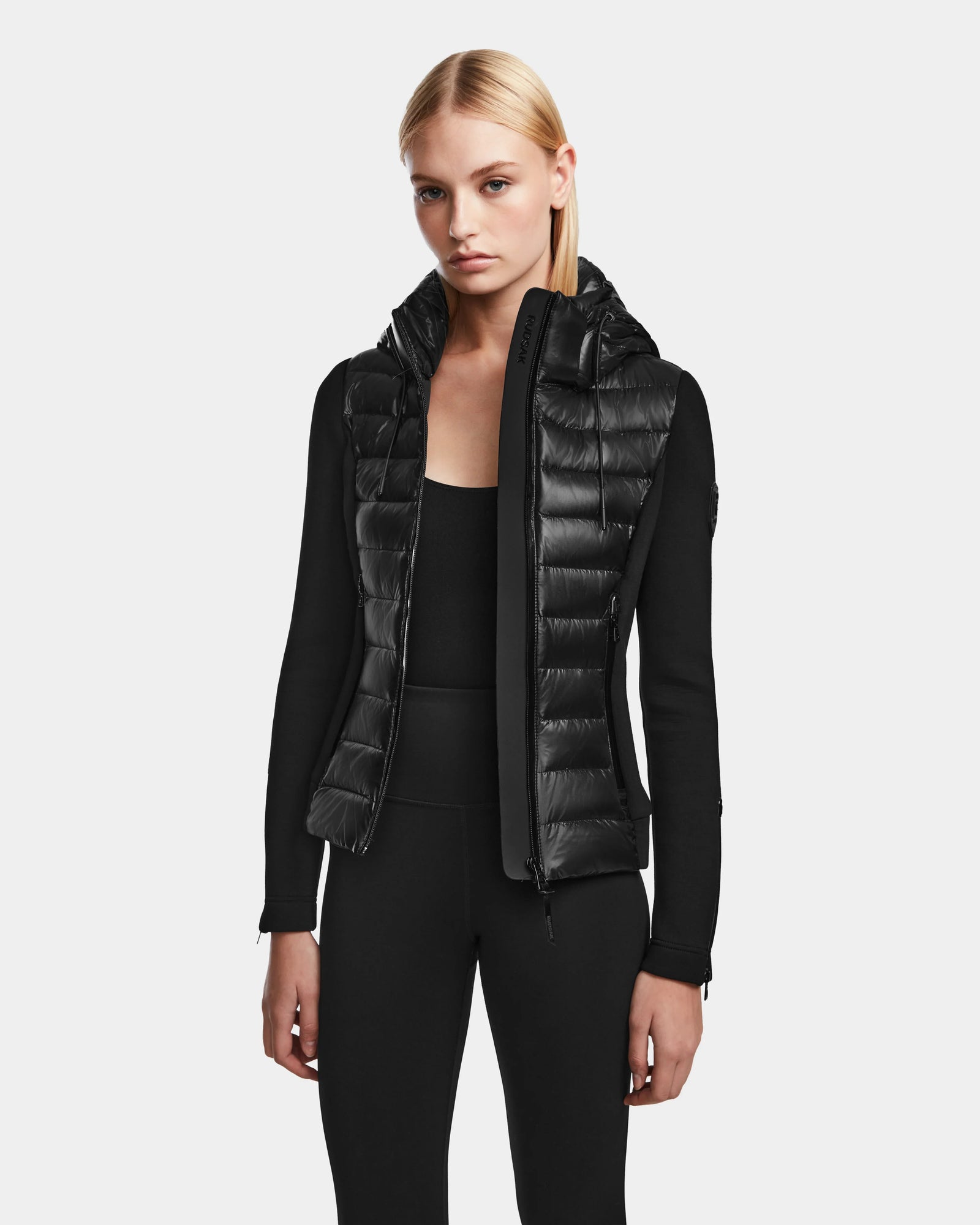 A woman with straight blonde hair models the RUDSAK MABS - 8122020 BLACK women’s puffer jacket over a black long-sleeve top and pants, standing against a plain light gray background.
