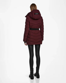 A person stands facing away, wearing the Rudsak MALAYA M - L123554 Wine womens down puffer jacket with herringbone quilting and hood, styled with a black belt, leggings, and boots against a plain white background.