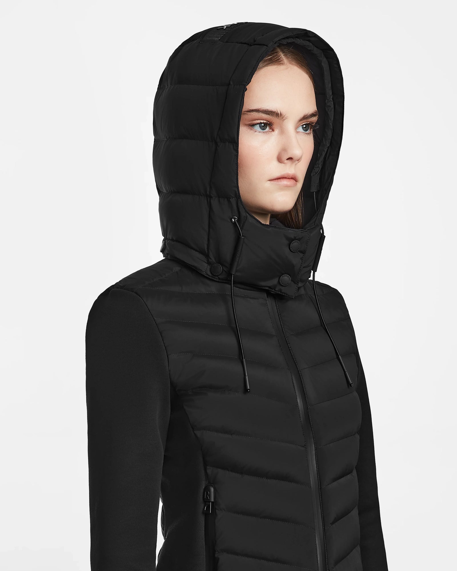 A young woman wears the Rudsak MALYA M - L123547 Black puffer-style hooded jacket with a front zipper and drawstring hood, looking forward with a neutral expression against a plain white background.