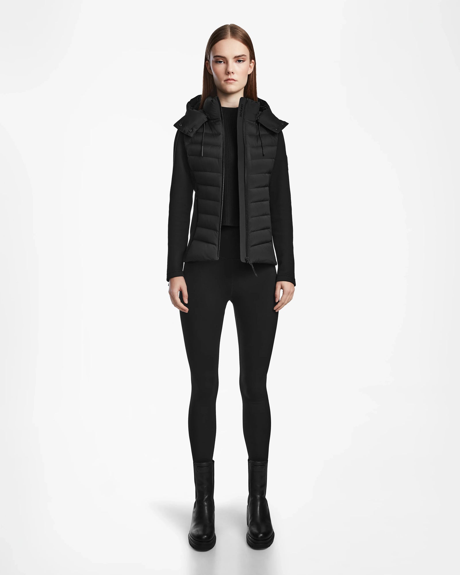 A woman faces forward in a Rudsak MALYA M - L123547 Black down quilted jacket with hood, black long-sleeve top, leggings, and ankle boots, set against a plain white background.
