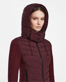 A woman models the Rudsak MALYA M - L123547 Wine jacket, a maroon down quilted style with hood, black zippers, and drawstrings—perfect as a cozy lightweight jacket for women.