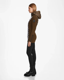 A woman with straight blonde hair stands in profile wearing the Rudsak MARRY - L121003 Light Olive puffer, black leggings, and black ankle boots against a plain white background.