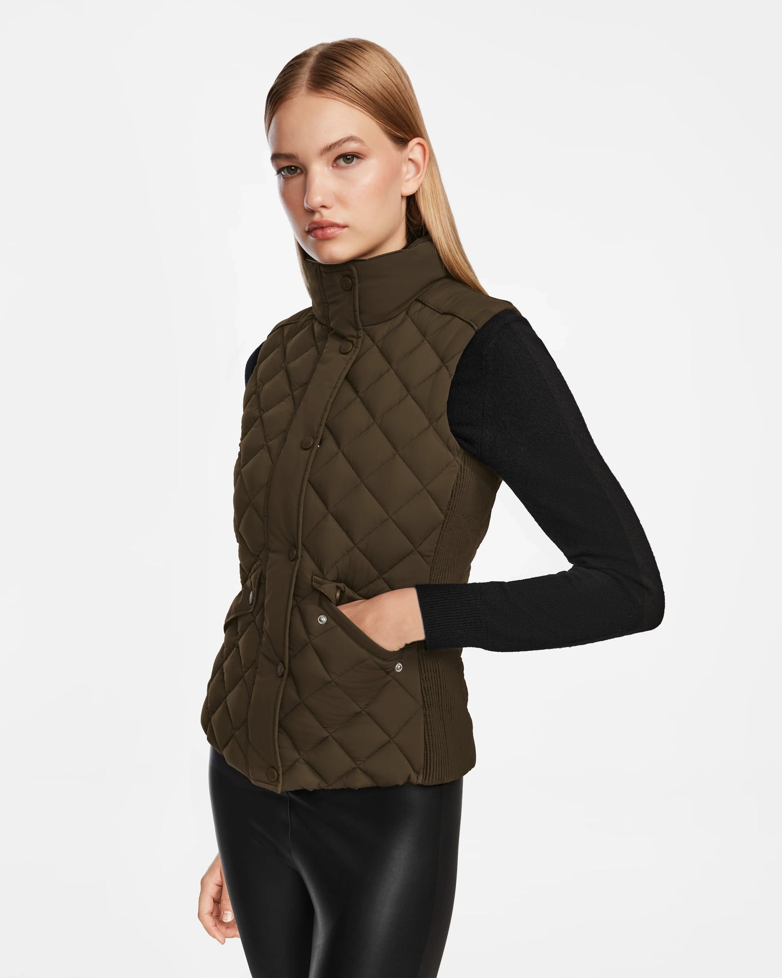 A woman with straight blonde hair wears a Rudsak MATILDA - L124504 Army diamond-quilted vest over a black long-sleeve top and black leather pants, posing with one hand in her pocket against a plain white background.