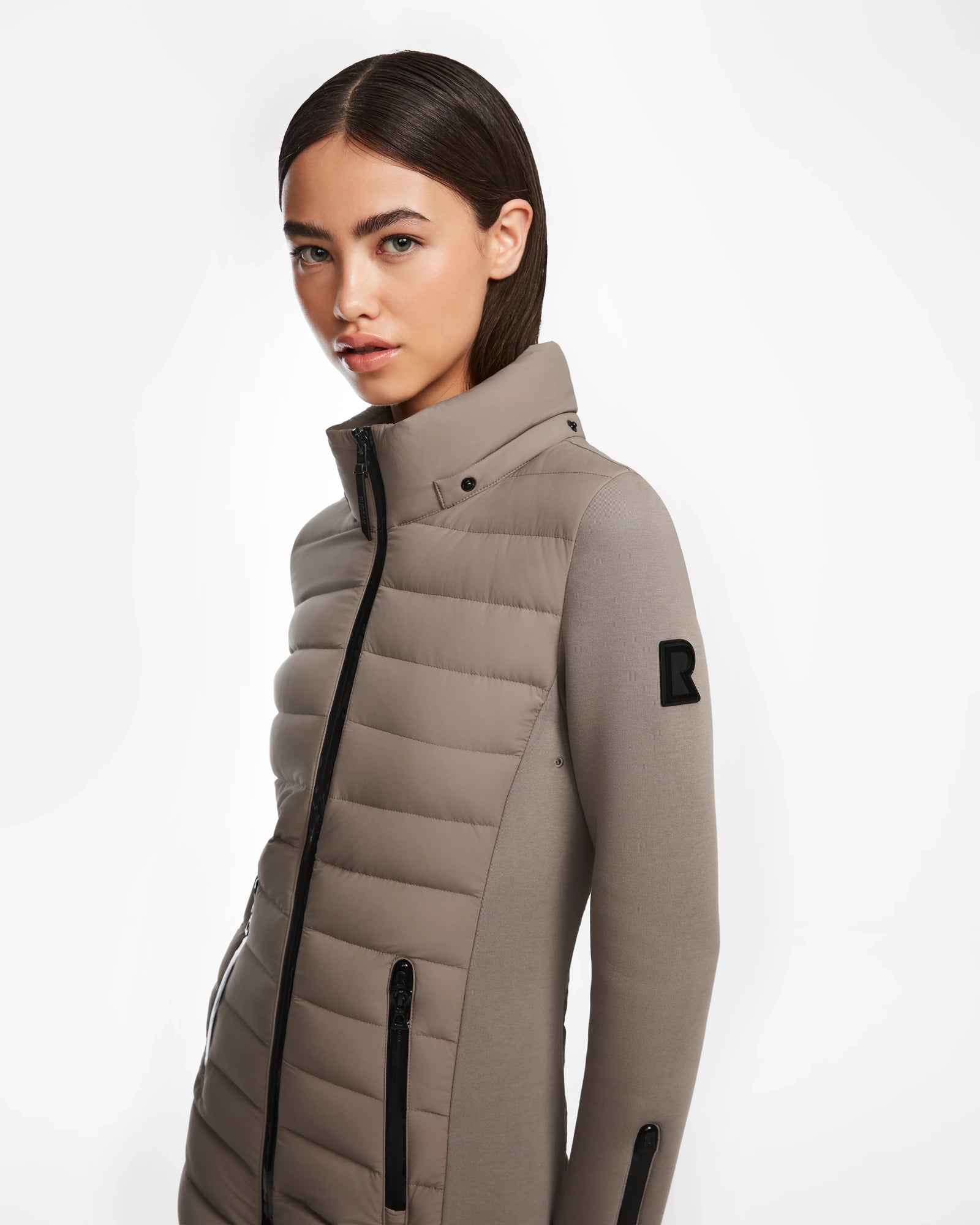 A woman with straight brown hair wears the Rudsak MERINA M - 8124030 Cement women’s light taupe jacket, padded with zippered pockets, high collar, and a black R patch on the sleeve, standing against a plain white background.