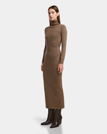 A woman stands sideways in a body-hugging, ankle-length MONICA - 8524516 Antique Bronze knit dress by Rudsak with long sleeves and a turtleneck, paired with dark brown heeled boots, against a plain white background.