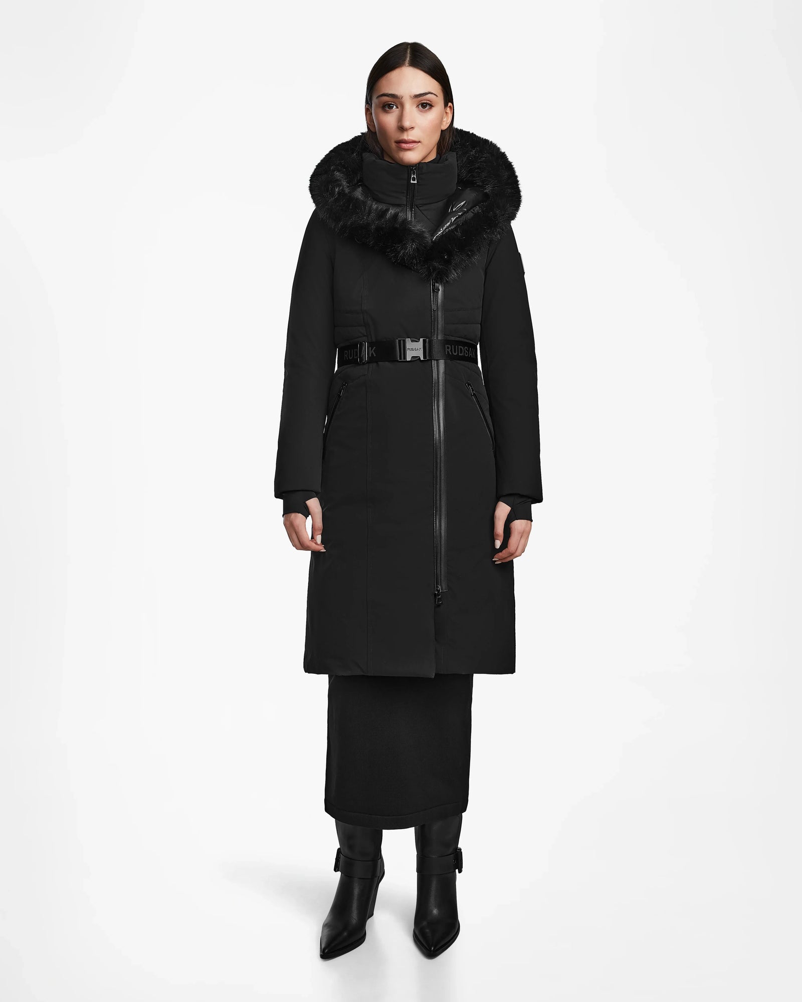 A woman with dark hair faces forward wearing the Rudsak MUSA - L124534 Black womens long down puffer, featuring a belted waist and a large black fur-trimmed hood, paired with a long black skirt and black heeled boots.