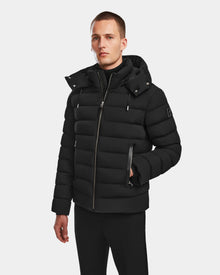 A man in the Rudsak NAVAN - 6124551 Black mens quilted down puffer with a high collar and hood stands against a white background. The RDS-certified jacket is wind and water-repellent. He has short hair, a neutral expression, and one hand in his pocket.