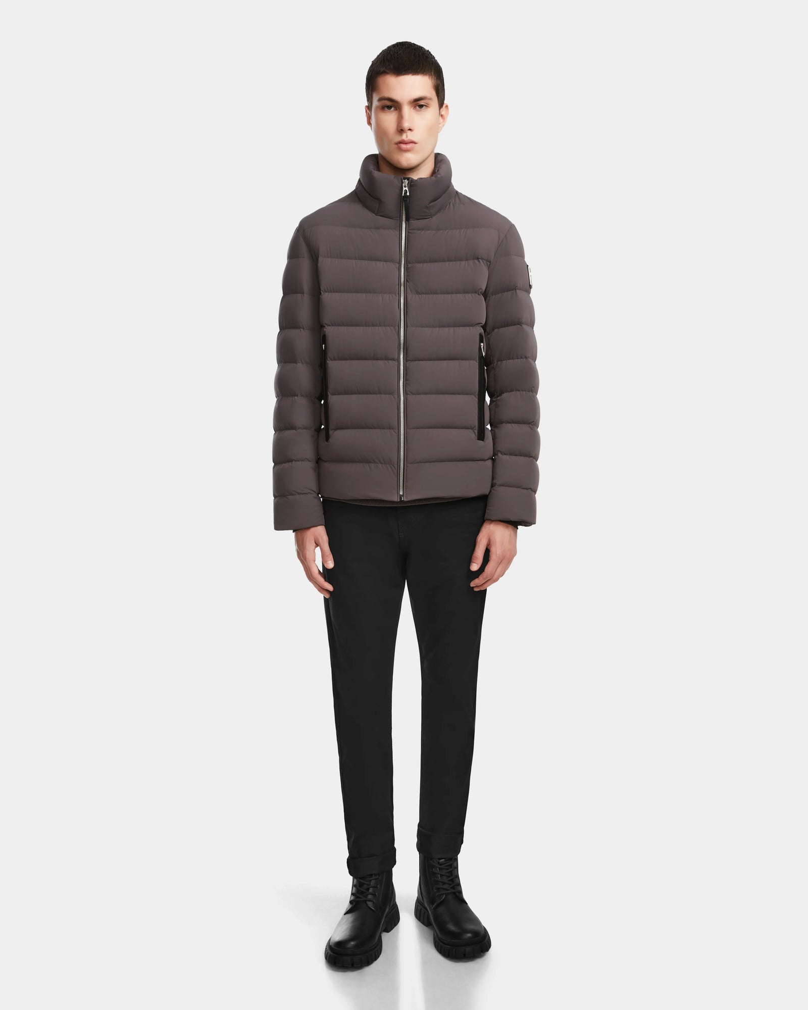 A man stands facing forward, wearing the Rudsak NAVAN - 6124551 Grey quilted down puffer, black pants, and black boots against a plain white background.