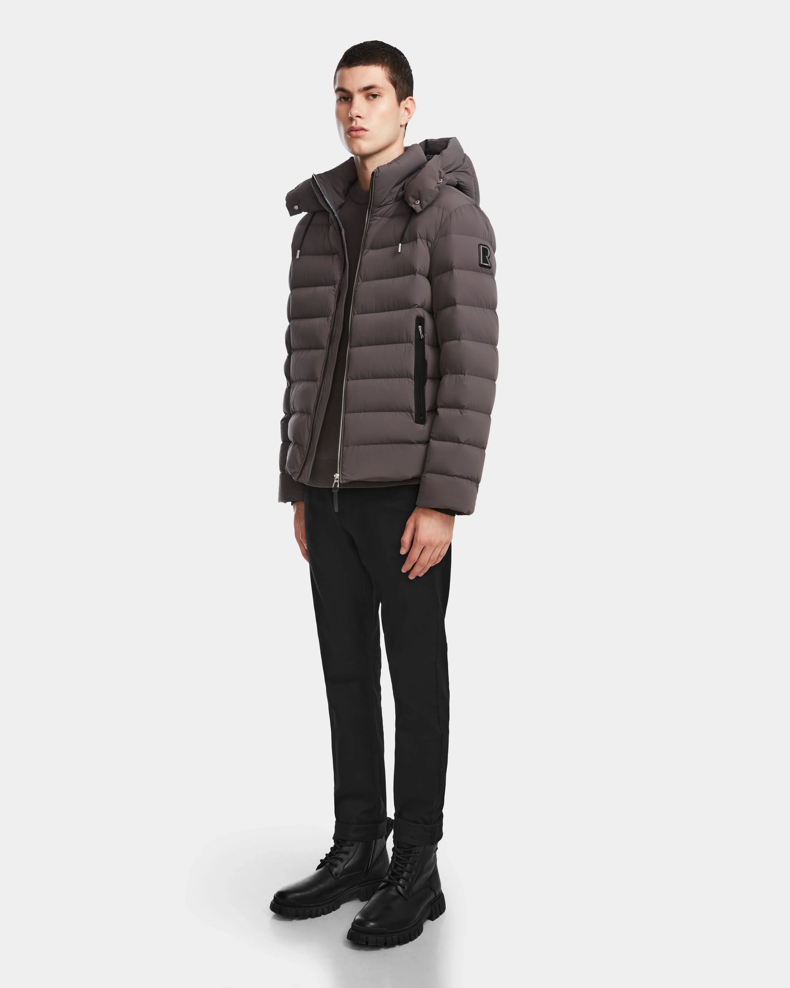 A young man stands facing forward, wearing the Rudsak NAVAN - 6124551 Grey mens quilted down puffer jacket with RDS-certified down, paired with black pants and black boots, against a plain white background.